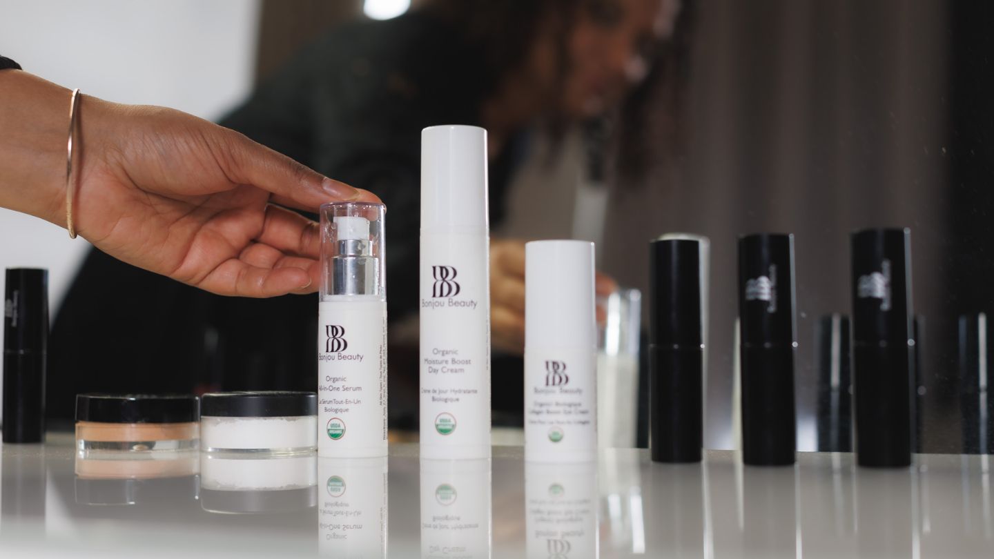 woman holding skincare product bottles on a table with her reflection in a mirror
