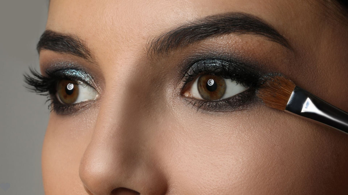 Close up of a woman's eyes with smoky eye makeup and a brush on a grey background