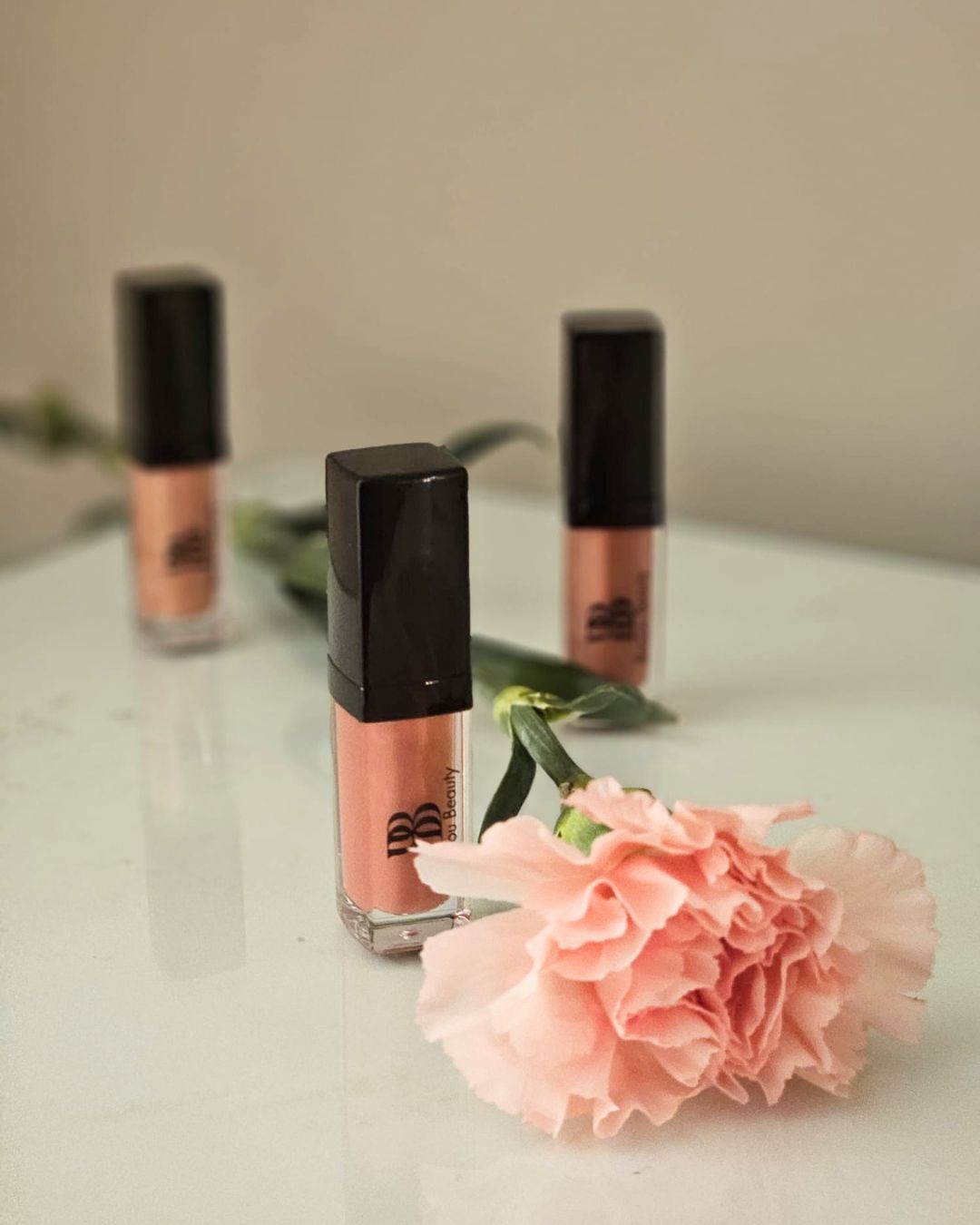 Three bottles of Bonjou Beauty pinky-peach  lip gloss with black caps arranged on a white surface with a pink carnation flower in the foreground.