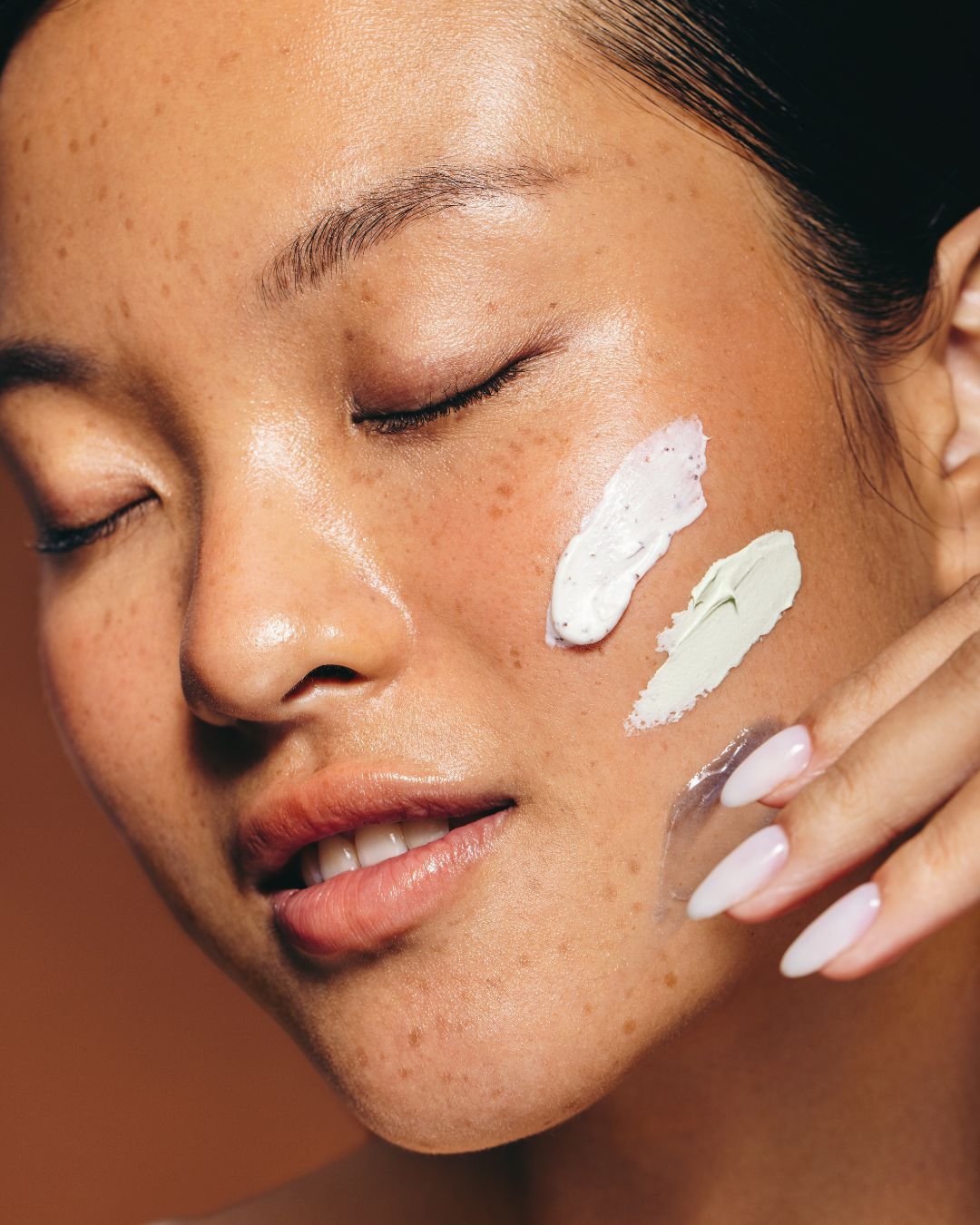 Close-up of a woman's face with moisturizer swatches applied to her cheek against a warm brown background.