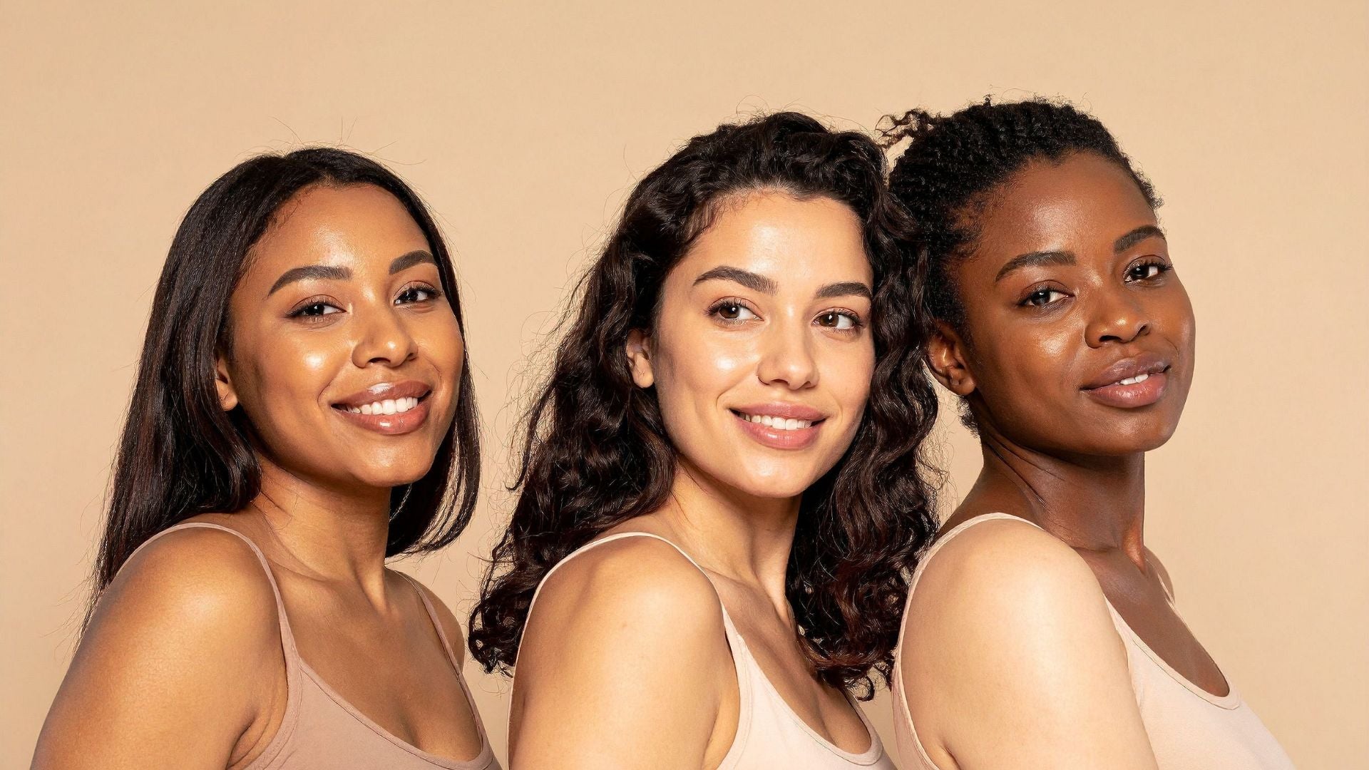 Three women with different skin tones and hair textures posing together against a solid beige background, wearing matching beige tank tops.