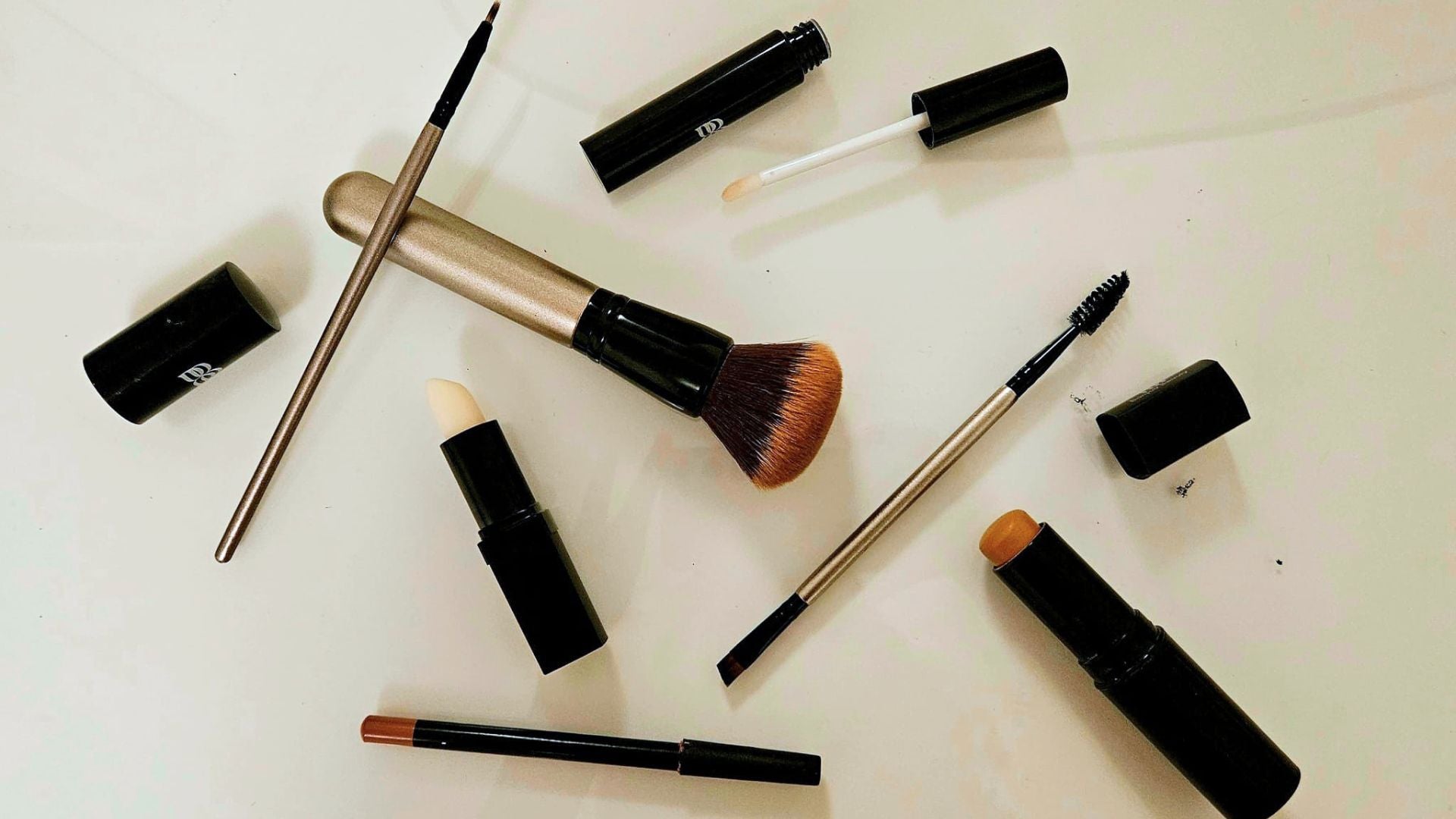 An overhead flat-lay of several  Bonjou Beauty makeup items on a plain white surface, including a makeup brush with a beige and black handle, a lip product tube, a small applicator wand, a pencil, and other black cosmetic caps.