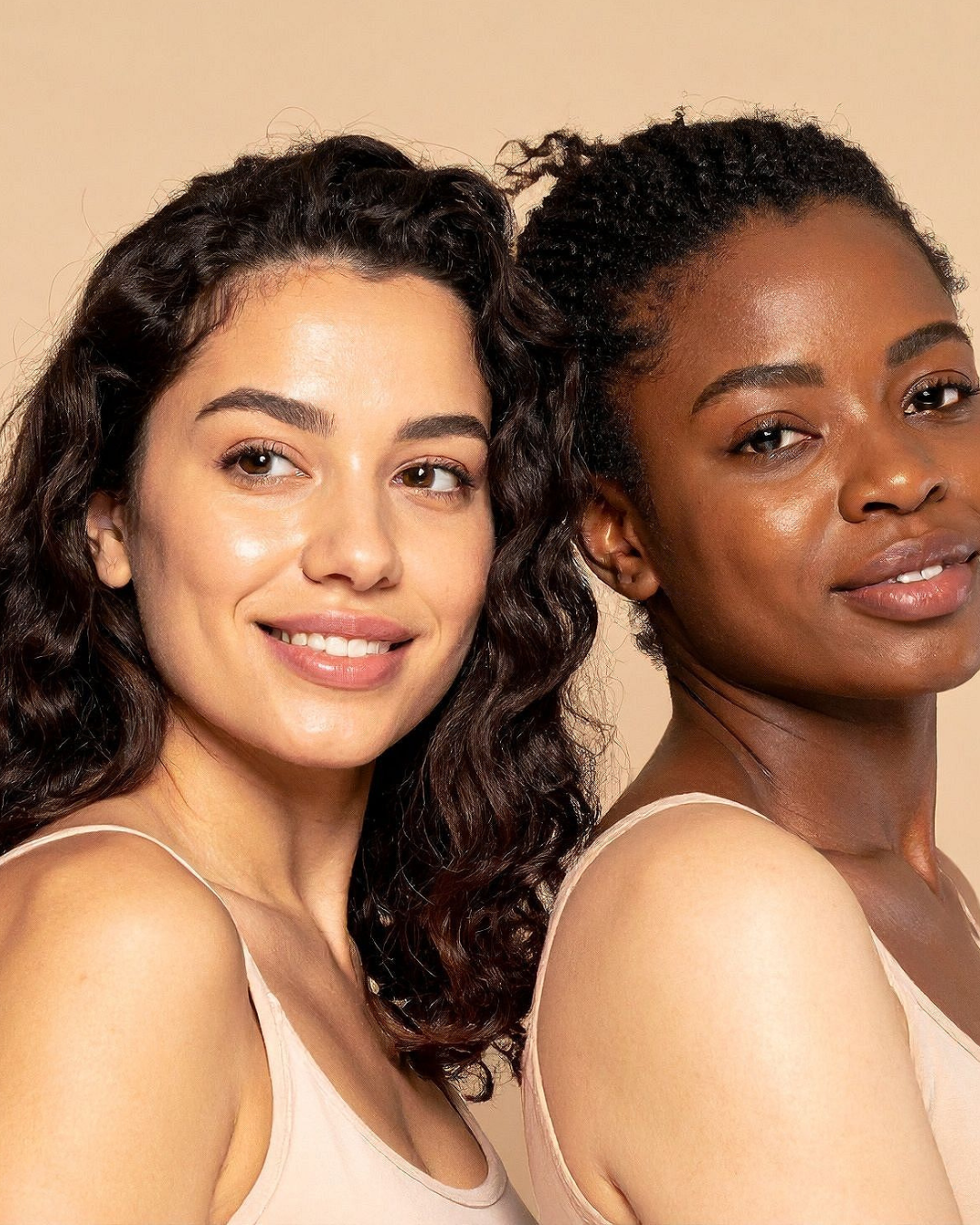 Two women with dark hair and different skin tones wearing matching beige ribbed tank tops, posed against a solid beige background.