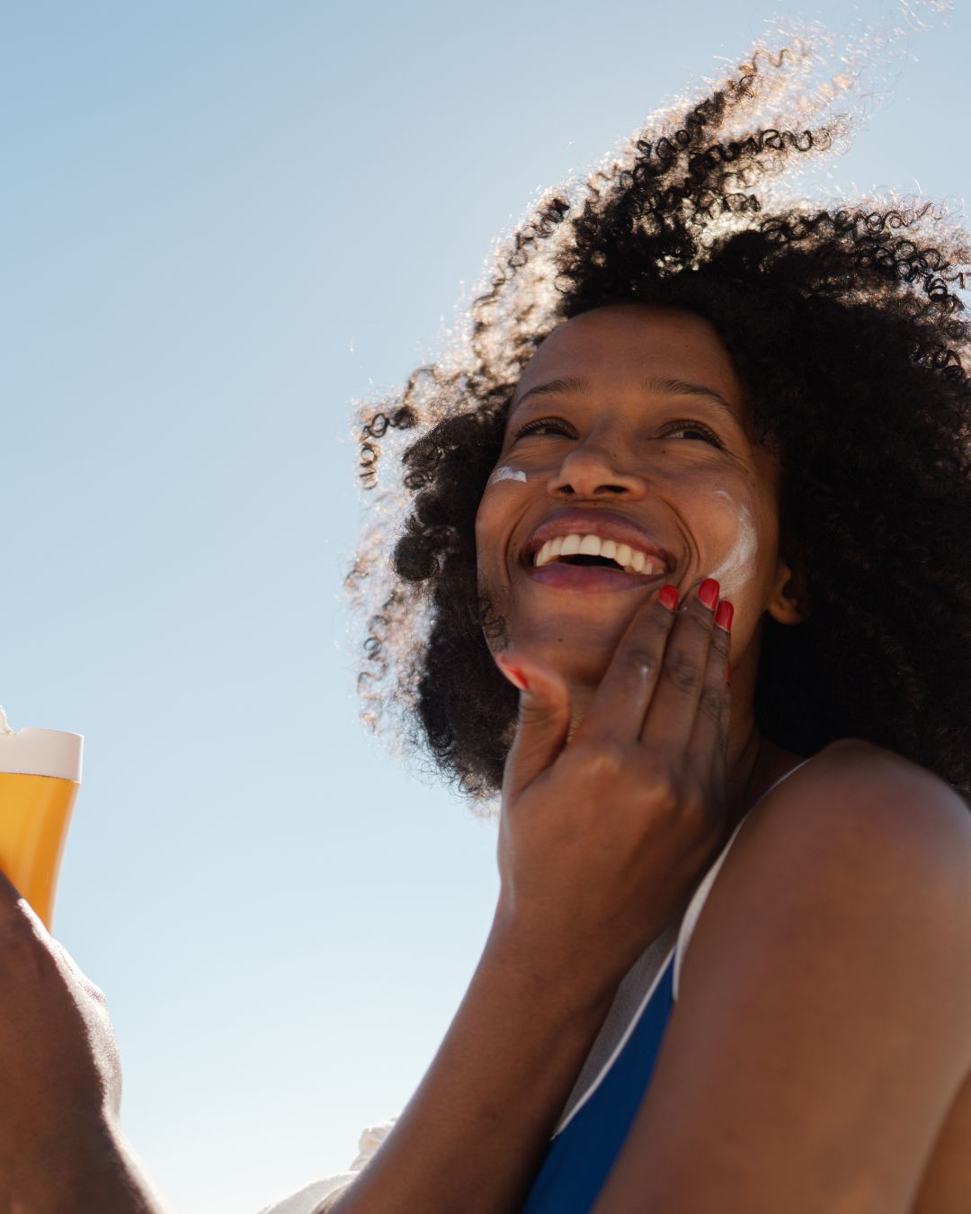 A smiling woman with curly hair applies sunscreen cream to her face outdoors under a clear blue sky, holding a yellow drink in one hand.