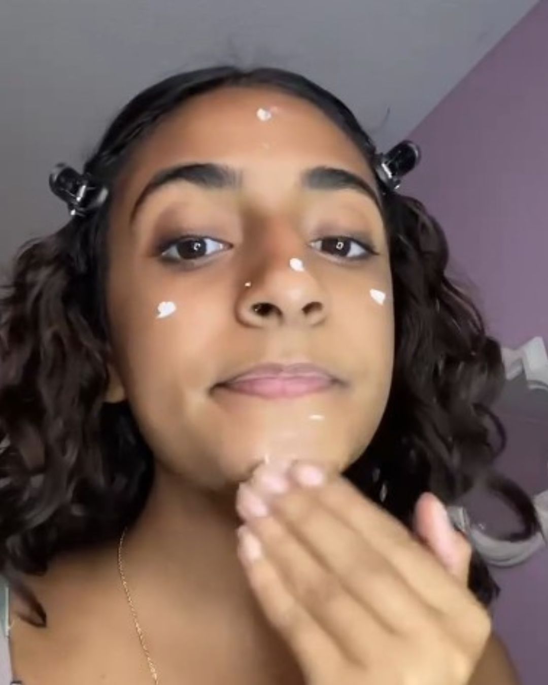 A close-up selfie of my niece with dark curly hair, showing white cream being applied to her face and hand, with clips holding her hair in place.