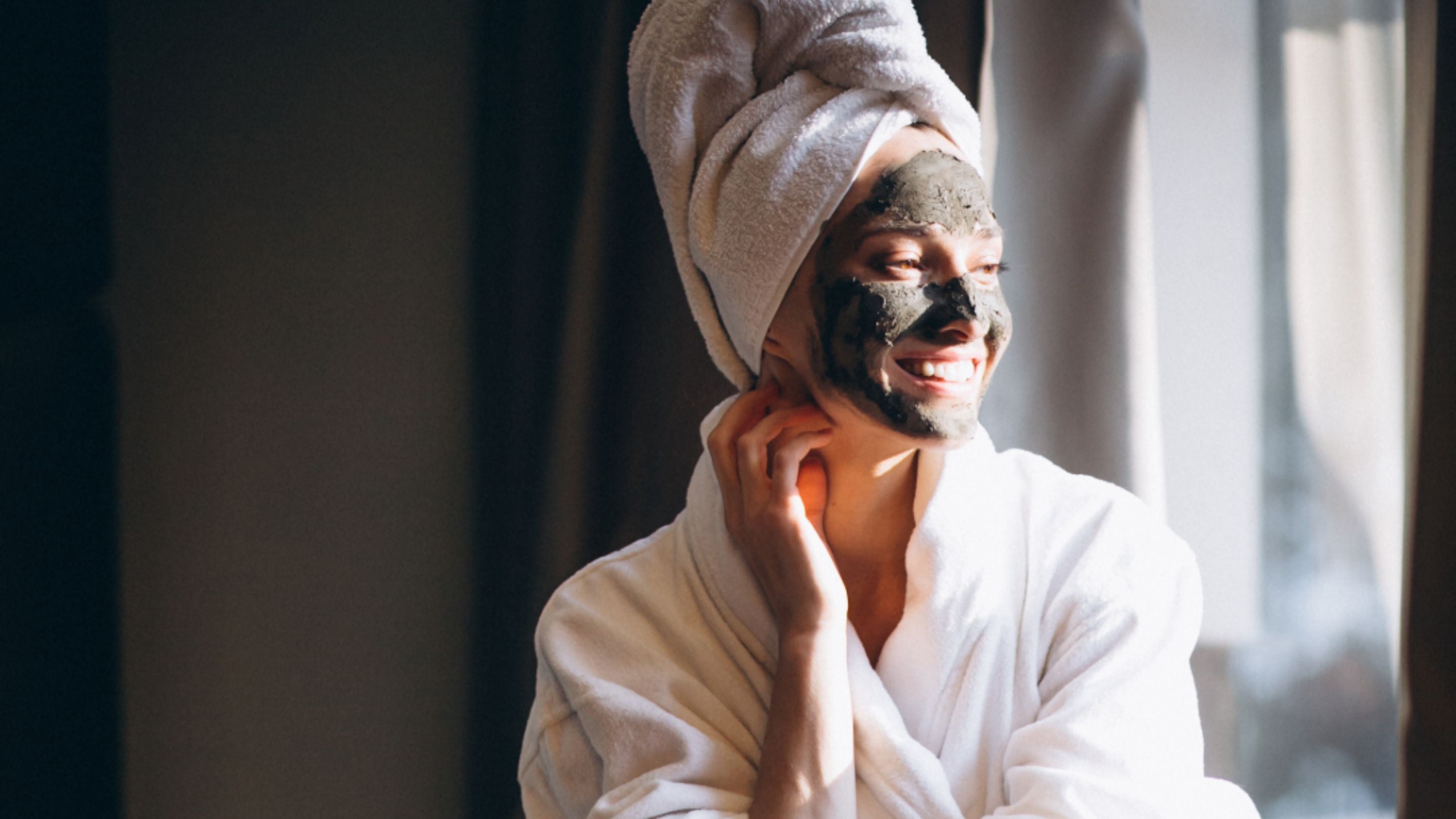 Woman in a white robe with a towel on her head, with a mineral mud face mask sitting by a window.