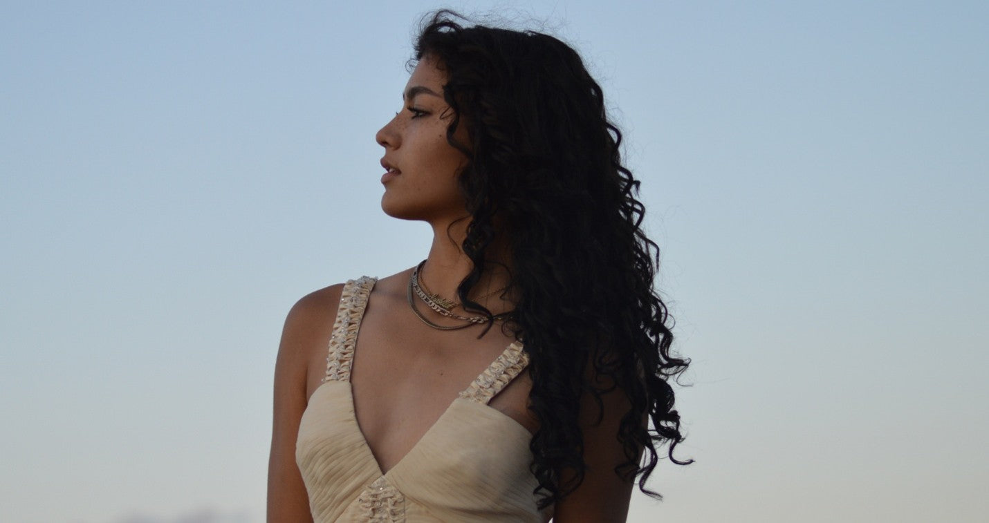 My niece with long, curly black hair is shown in profile against a pale blue sky, wearing a cream-colored lace-trimmed top and layered necklaces.