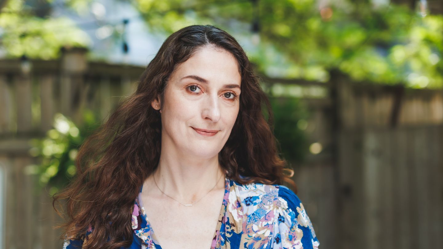 A woman with long wavy brown hair, natural makeup, wearing a blue floral-patterned top, photographed outdoors against a blurred wooden fence and green foliage background.