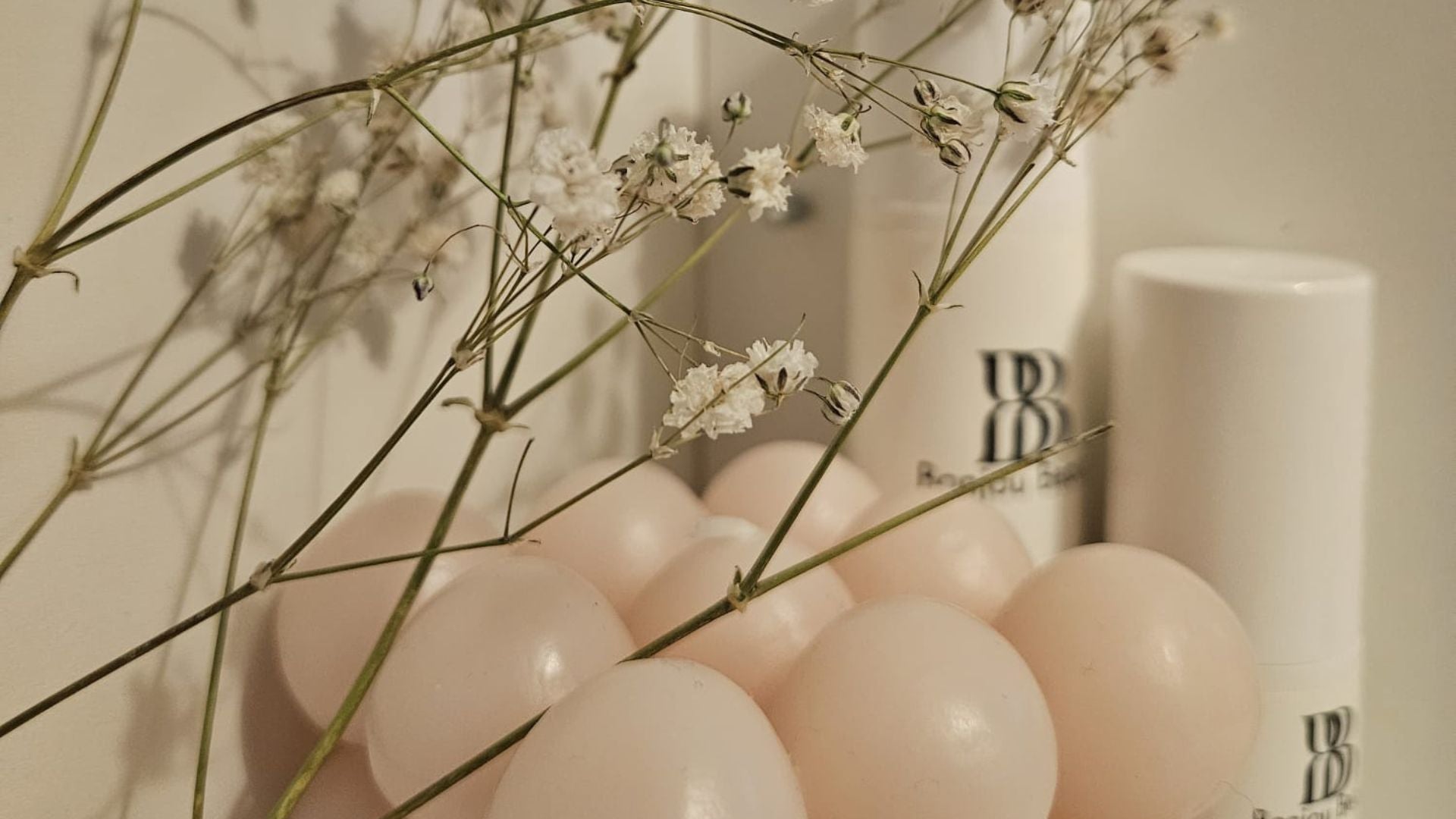 White cosmetic bottles and a white stick container with a black 'BB Bonjou Beauty' logo, styled with pale blush pink balloons and delicate white baby's breath flowers in the foreground.
