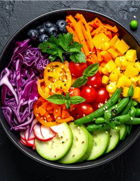A top-down view of a black bowl filled with a colorful, neatly arranged salad of vegetables including red cabbage, carrots, tomatoes, cucumbers, green beans, radishes, blueberries, and herbs on a dark textured background.